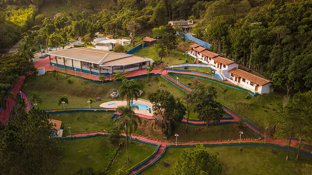 Vista aérea da propriedade com gramados, piscina, caminhos vermelhos e construções entre a mata